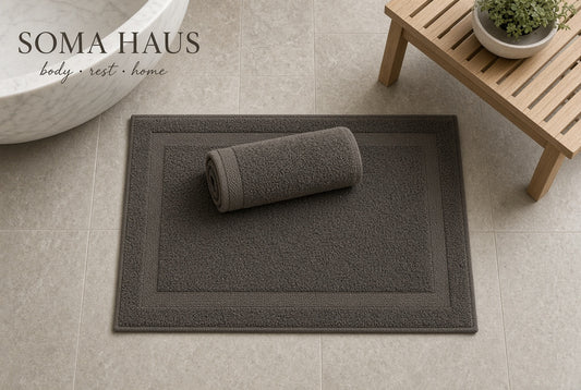 A dark gray rectangular bath mat with a matching rolled towel, shown on a light beige tiled bathroom floor. A wooden bench with a small potted plant and part of a white bathtub are visible in the background.