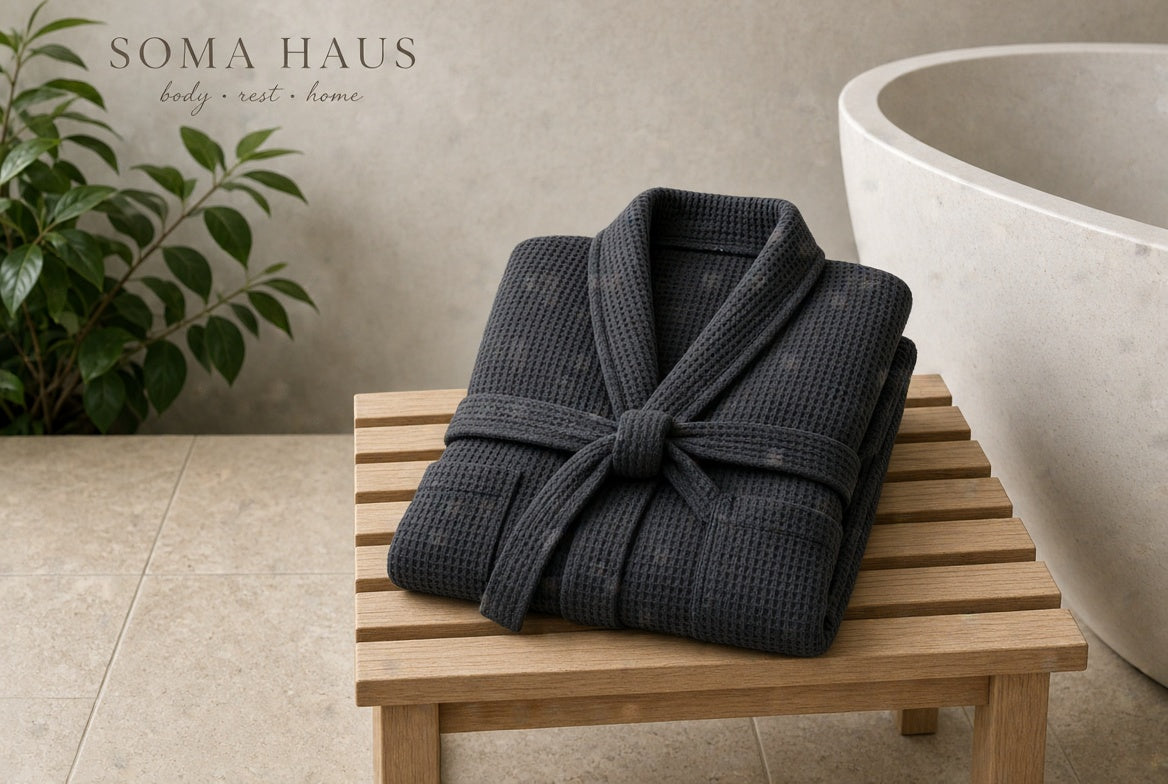 A dark charcoal gray waffle-weave bathrobe with a tie belt, neatly folded and displayed on a light wood slatted stool in a bathroom setting. A white bathtub is visible on the right, a green plant on the left, and the brand name 'SOMA HAUS' is printed in the upper left corner.