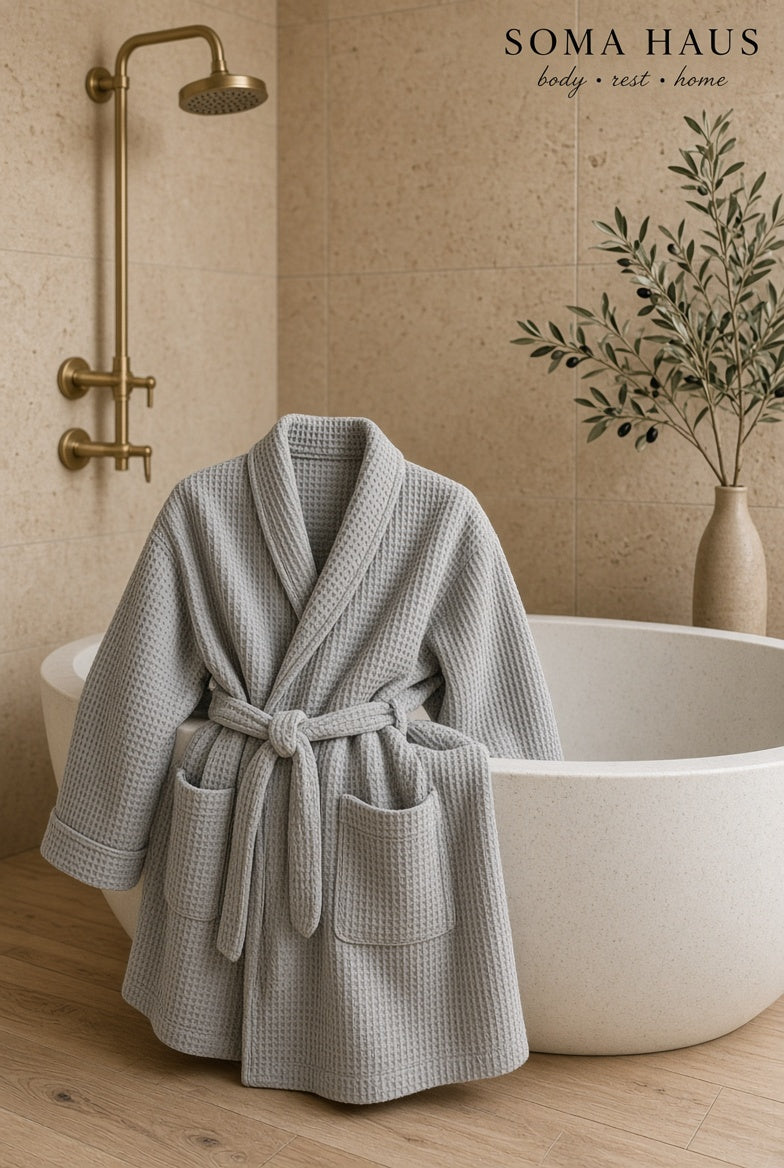 A light gray waffle-weave bathrobe with a tie belt and front pockets, draped over the edge of a white freestanding bathtub in a warm beige bathroom. A brass shower fixture is visible on the left, and a vase with dried leaves sits on the right.