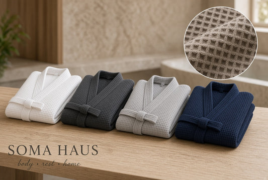 Four folded waffle-weave bath robes in white, charcoal gray, light gray, and navy blue are arranged on a wooden surface, with a circular inset showing a close-up of the waffle texture. The text 'SOMA HAUS body + rest + home' appears at the bottom left.