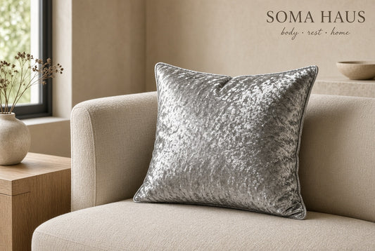 A square silver crushed-velvet throw pillow displayed on a beige fabric sofa in a warm, modern living room setting. The brand name 'SOMA HAUS' is visible in the top right corner.