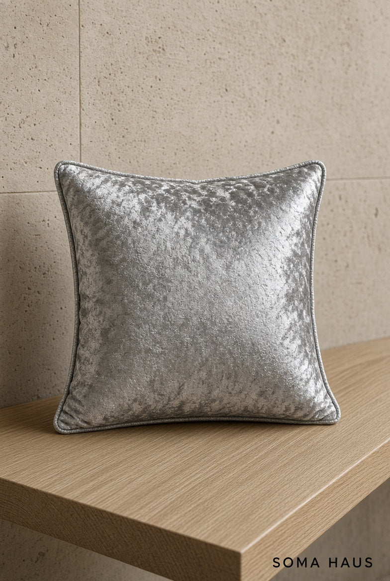A square silver-gray crushed velvet throw pillow with dark piping, resting on a light wood shelf against a beige stone wall. The brand name 'SOMA HAUS' is visible in the bottom right corner.