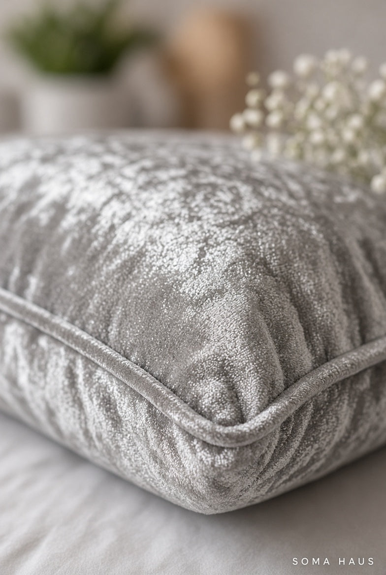 A close-up of a silver-gray crushed velvet throw pillow with a piped edge, resting on a light-colored surface with soft-focus decorative plants in the background.