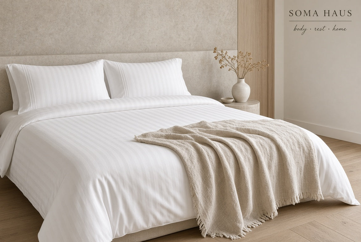 A neatly made bed in a minimalist bedroom setting, dressed in crisp white striped bedding with two pillows and a light beige throw blanket draped across the foot. A small side table with a vase of dried stems sits beside the bed.