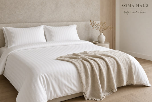 A neatly made bed in a minimalist bedroom setting, dressed in crisp white striped bedding with two pillows and a light beige throw blanket draped across the foot. A small side table with a vase of dried stems sits beside the bed.