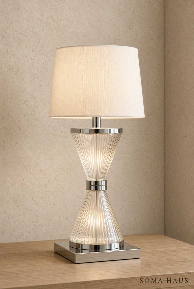 A modern table lamp with a ribbed clear glass hourglass-shaped base, polished silver metal accents, and a cream fabric drum shade, shown lit on a light wood surface against a textured beige wall. The brand name Soma Haus is visible in the bottom right corner.