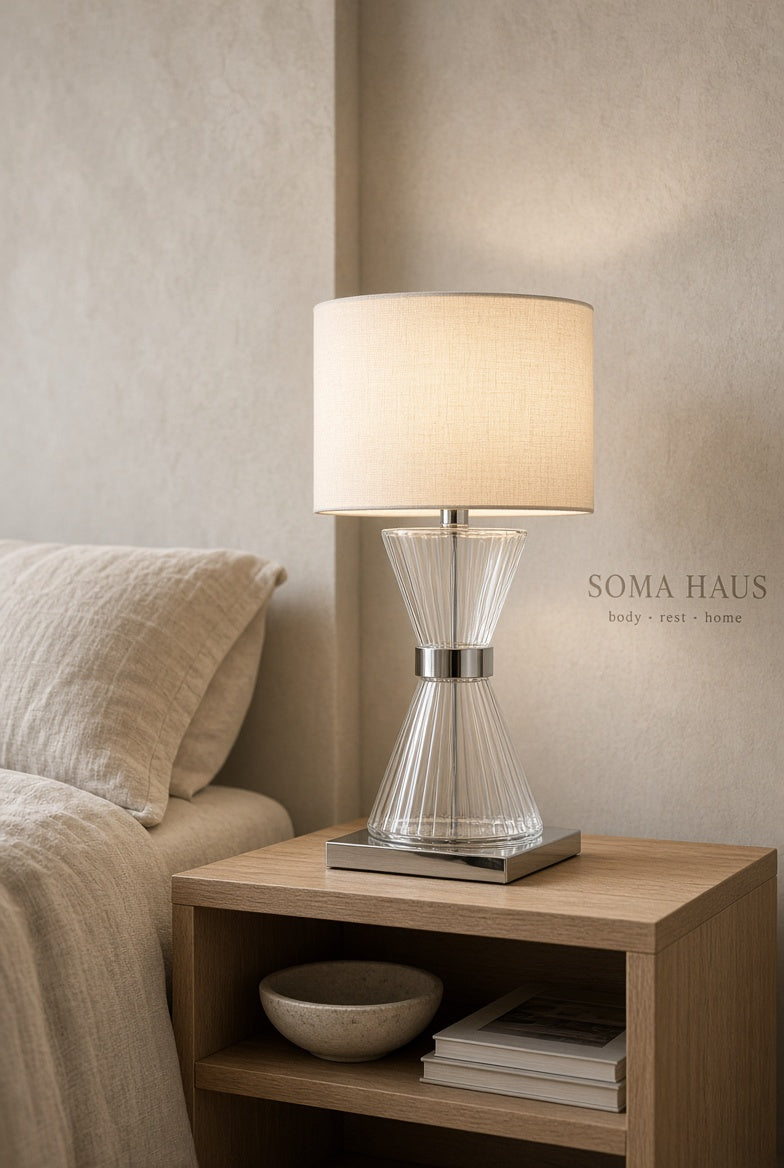 A Soma Haus table lamp with a clear ribbed glass base and a beige fabric drum shade, illuminated and styled on a light wood bedside table next to a bed.