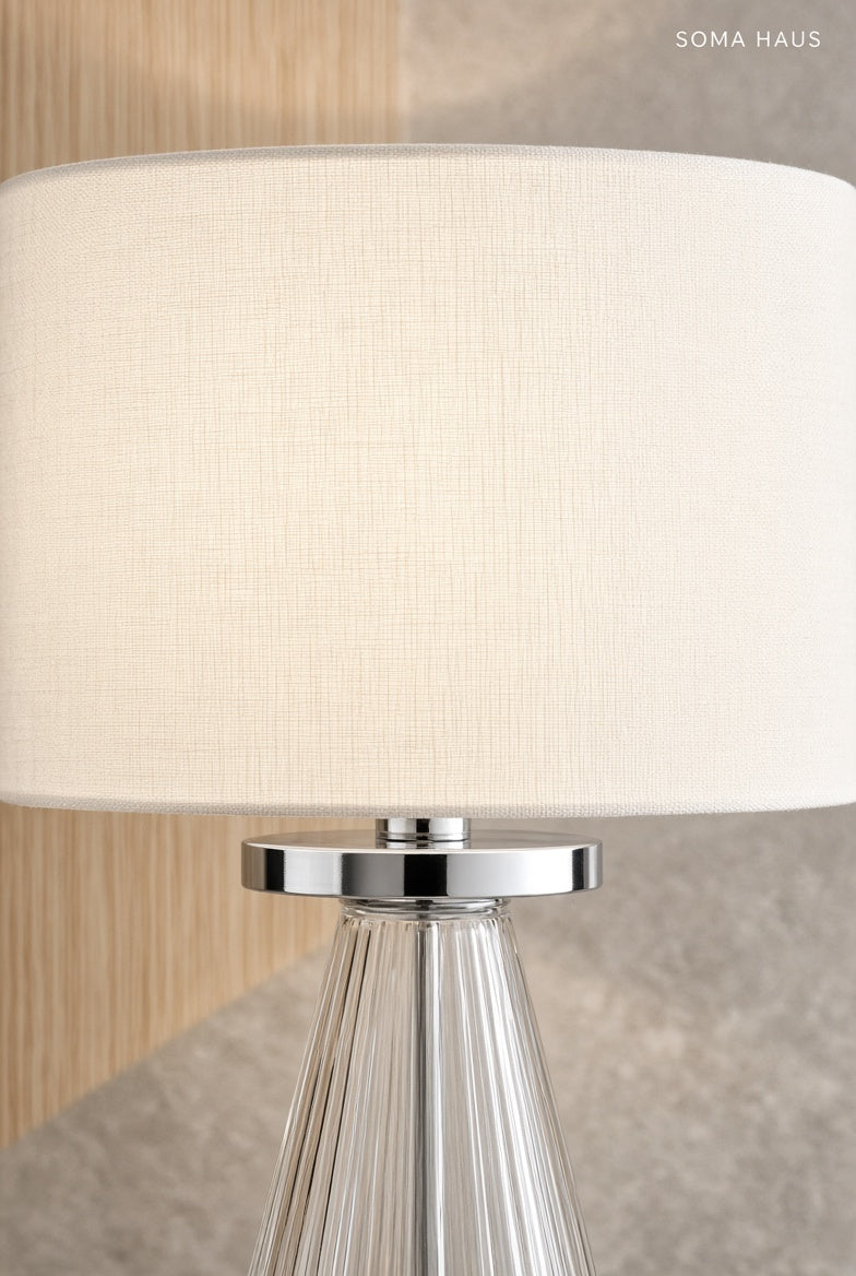 A close-up of a table lamp with a large off-white fabric drum shade, a polished silver metal collar, and a clear ribbed glass base. The brand name 'SOMA HAUS' is printed in the top right corner.