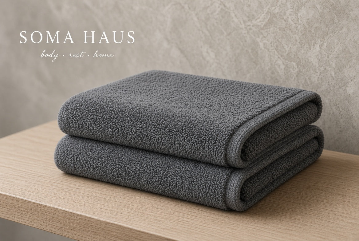 Two folded dark gray towels stacked on a light wood surface against a textured beige wall, with the text 'SOMA HAUS body rest home' in the upper left.