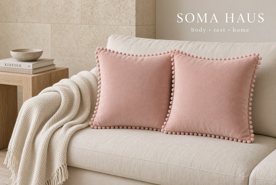 Two dusty pink velvet throw pillows with pom-pom trim resting on a cream sofa in a styled living room setting. A beige knit throw blanket is draped over the arm, and a small wooden side table with books and a bowl is visible on the left.