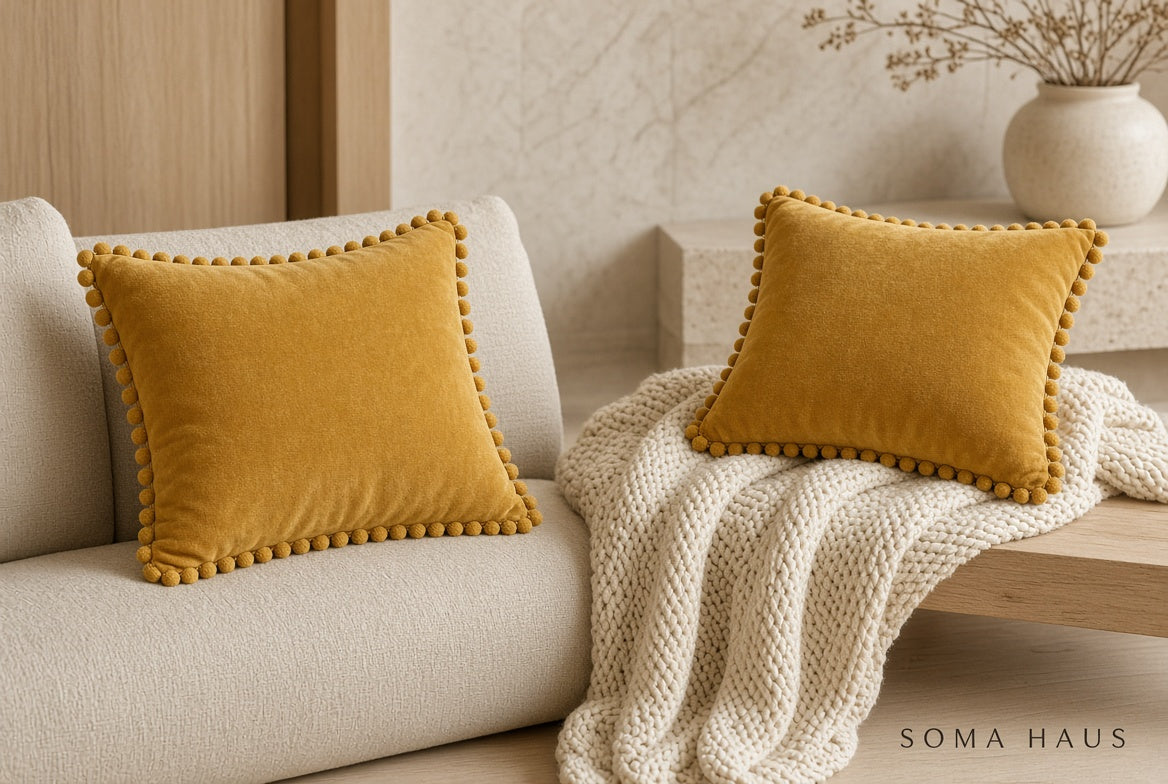 Two mustard yellow velvet throw pillows with pom-pom trim, styled on a cream sofa with a chunky knit cream throw blanket in a warm, modern living room setting. The brand name 'SOMA HAUS' is visible in the bottom right corner.