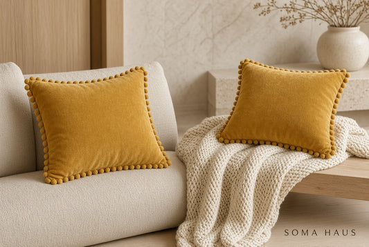 Two mustard yellow velvet throw pillows with pom-pom trim, styled on a cream sofa with a chunky knit cream throw blanket in a warm, modern living room setting. The brand name 'SOMA HAUS' is visible in the bottom right corner.