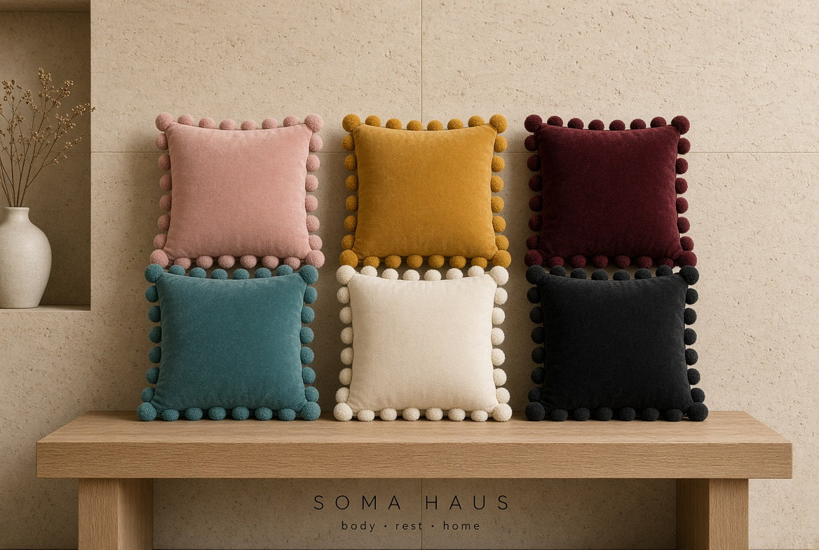 Six square decorative throw pillows with pom-pom trim arranged on a wooden bench against a beige textured wall. The pillows come in pink, mustard yellow, burgundy, teal, cream, and black.