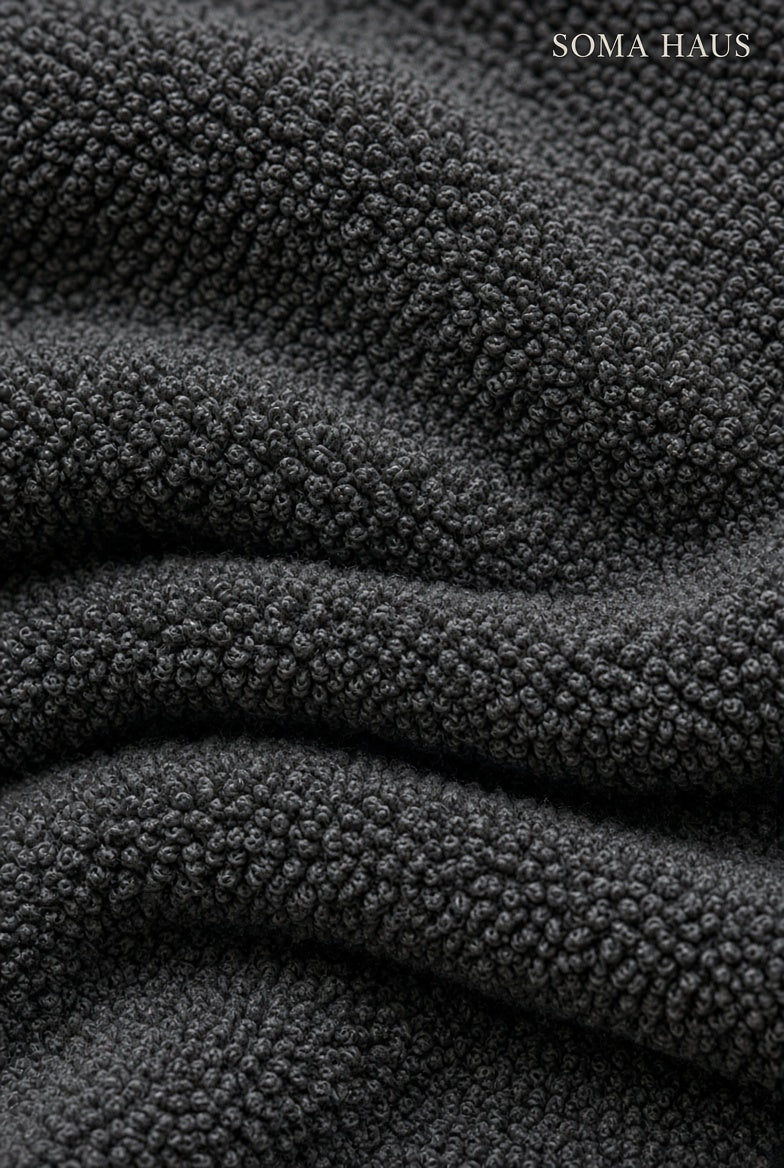 Close-up of dark charcoal gray textured knit fabric with soft folds and the brand name SOMA HAUS in the top right corner.