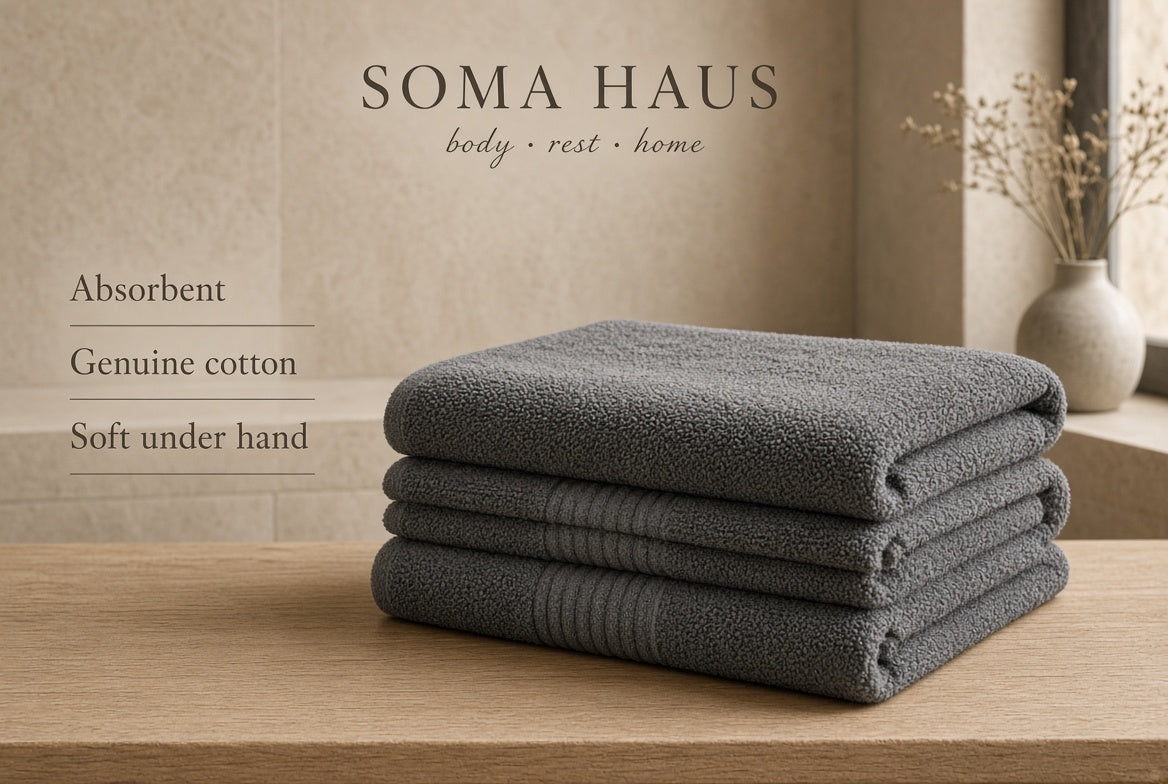 Three folded dark gray cotton towels stacked on a light wood surface, with the brand name Soma Haus and marketing text 'Absorbent Genuine cotton Soft under hand' on a beige wall, plus a vase with dried stems in the background.